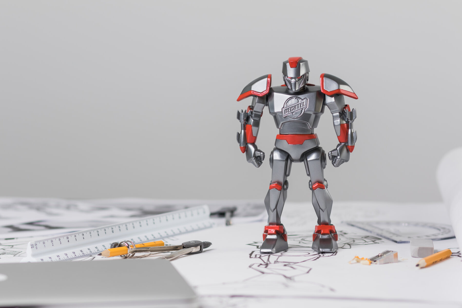 The BechMech toy, a silver and red armored robot, standing on a desk of blueprints beside a ruler and pencils.