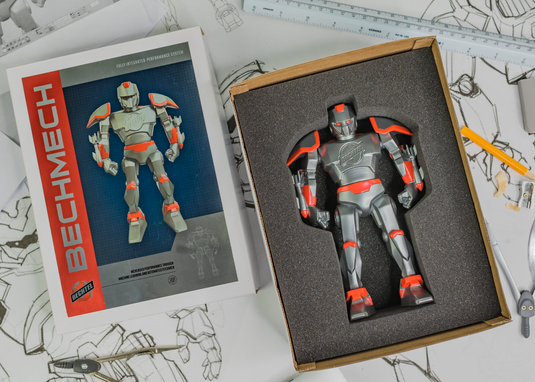 Overhead: BechMech in its foam tray beside the red BECHMECH box, on robot sketches.