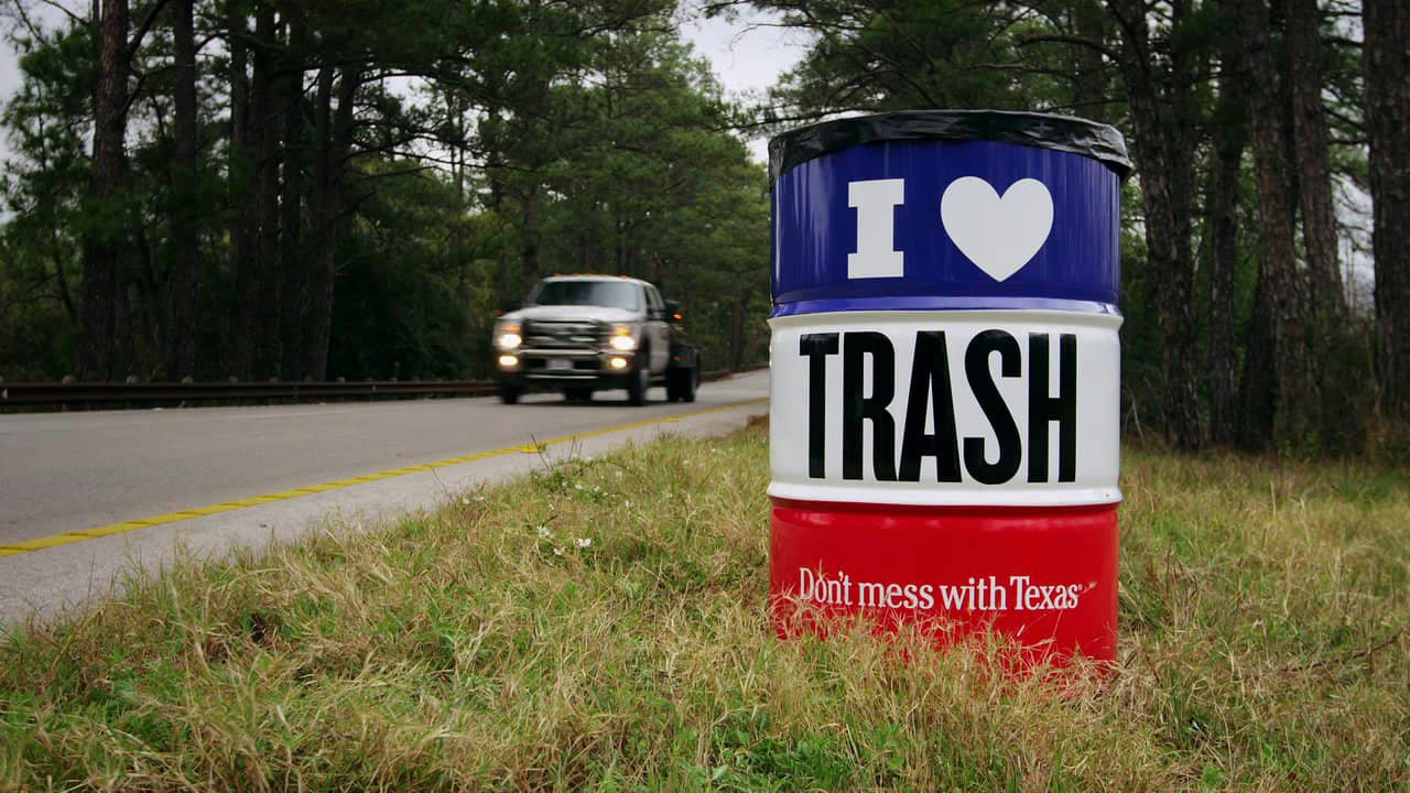 A red, white and blue barrel reading I love trash with the Don't mess with Texas script, beside a Texas highway as a pickup truck passes.