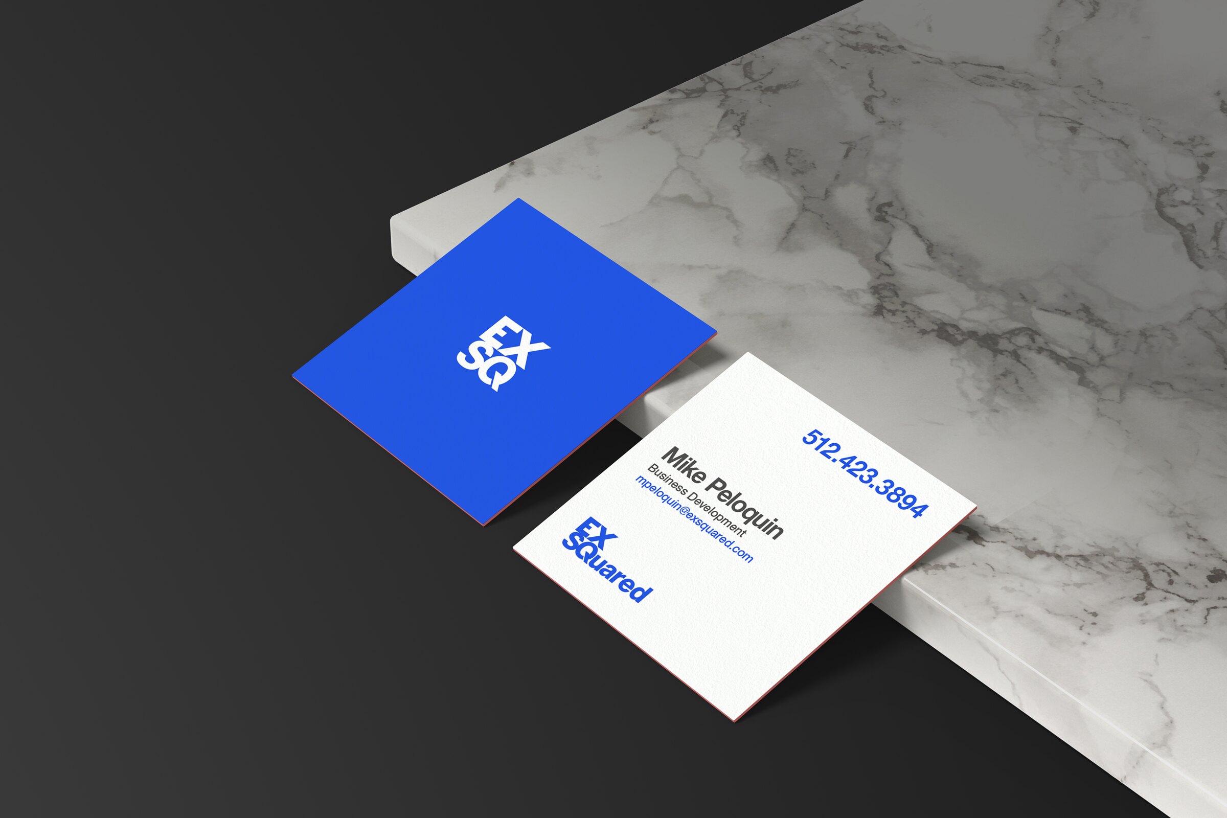 Two square EXSquared business cards on a marble ledge: the True Blue EXSQ mark on the front, the name, number and wordmark on the back
